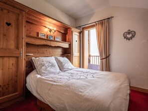 A bedroom with a wooden headboard, a bed with white bedding, a window with curtains, and a wall-mounted light fixture.