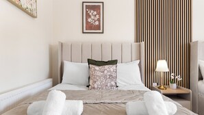 A neatly made bed with white towels, a floral pillow, and a bedside table with a lamp and a vase of flowers. - Stunning 2B Flat in South Kensington Near Station (London)