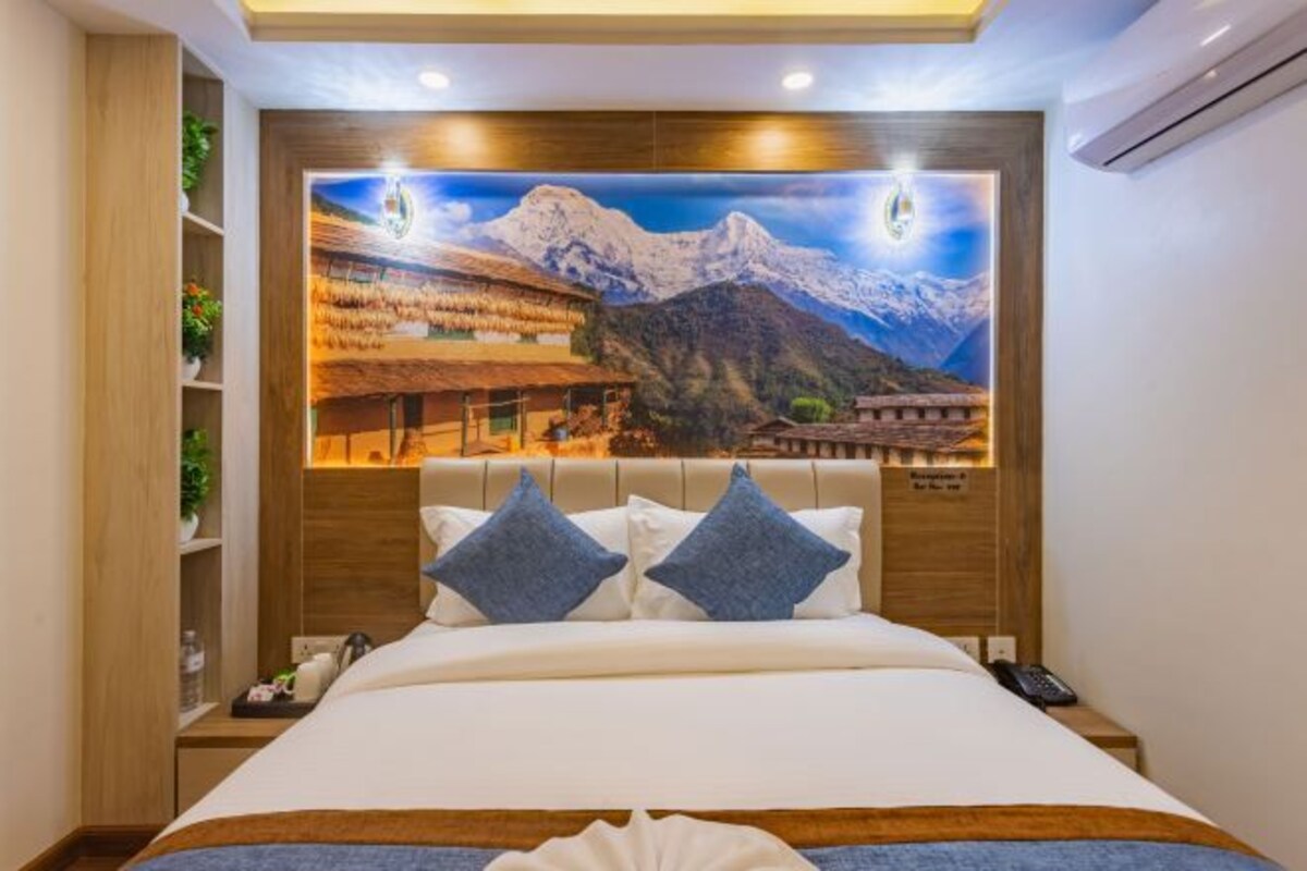 A hotel room with a large bed, a nightstand, a wall-mounted TV displaying a mountainous landscape, and a wooden headboard.