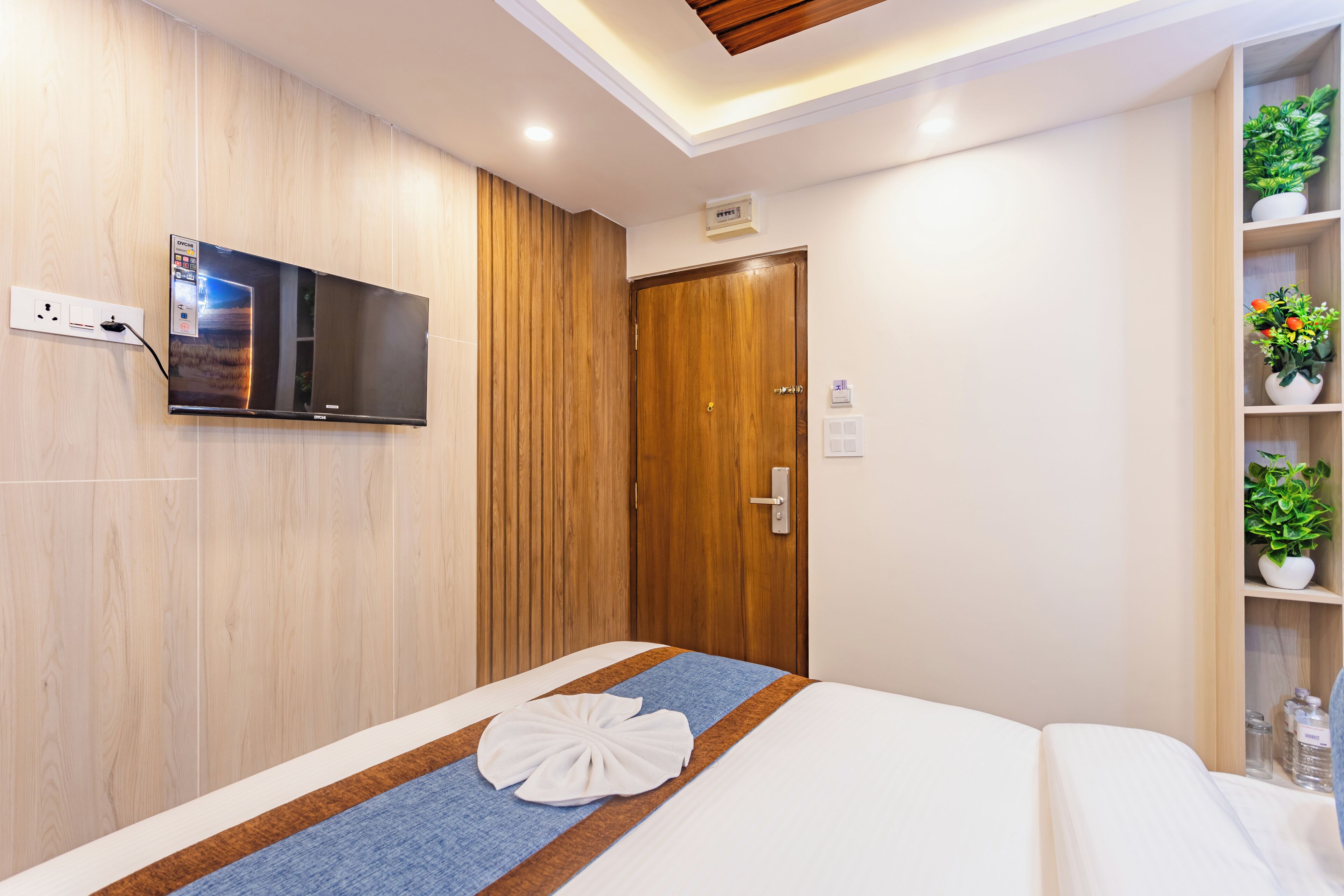 A modern hotel room with a wooden wall, a flat-screen TV mounted on the wall, a wooden door, a bedside table with a plant, and a neatly made bed with a towel on the bed.