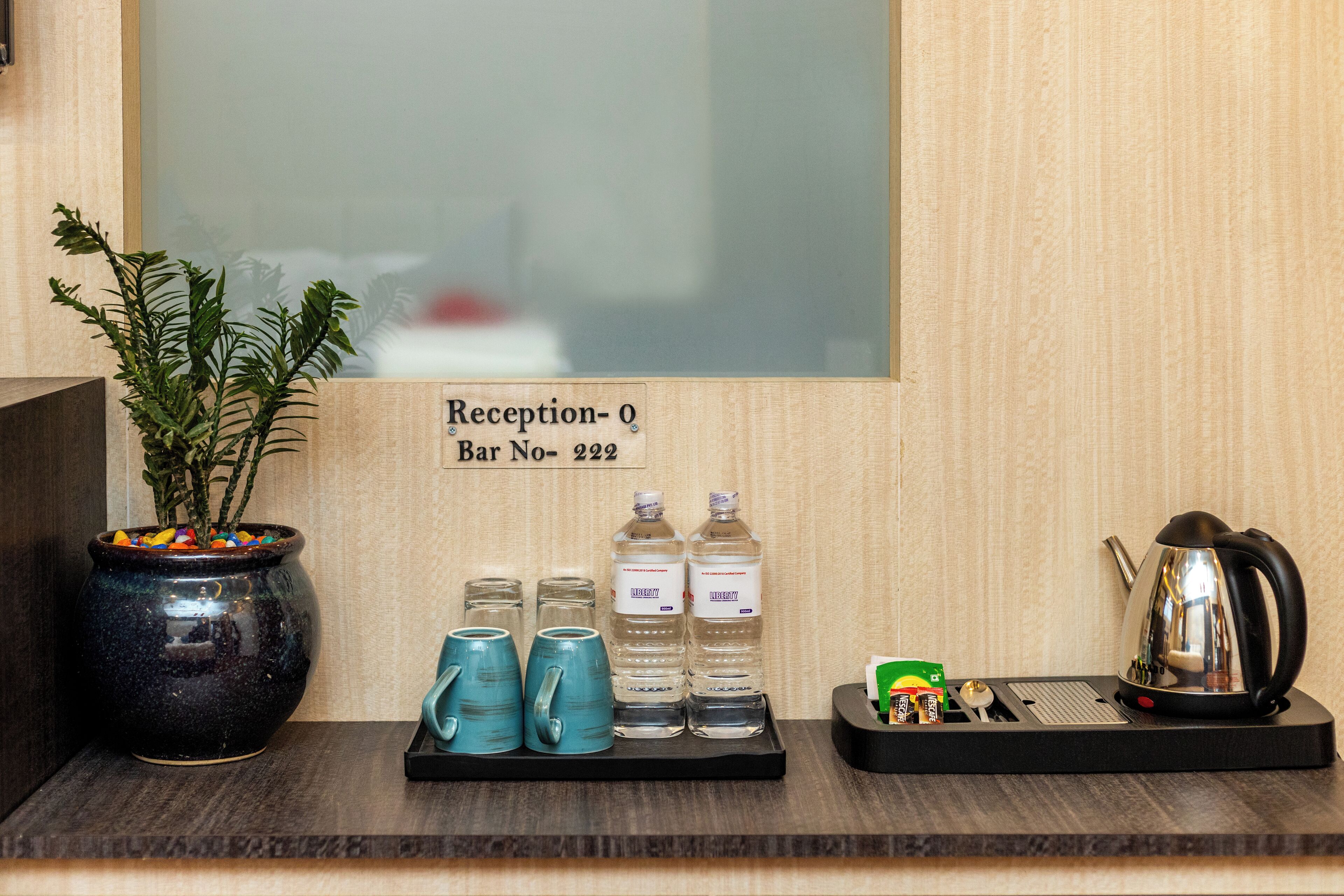 A reception area with a sign indicating "Reception-0 Bar No-222". There are two blue mugs, two water bottles, a small plant in a black pot, and a kettle on a tray.