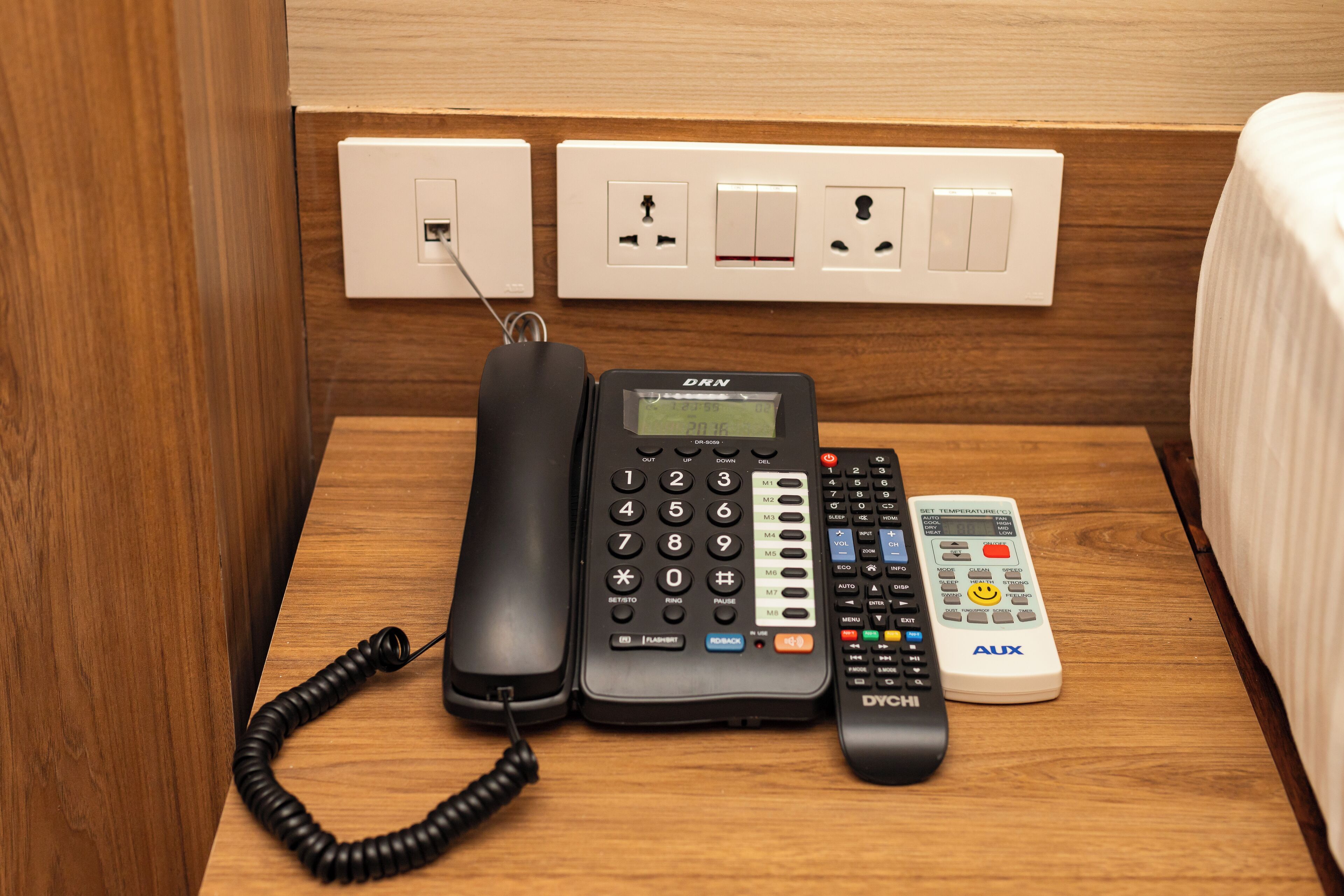 Una habitación de hotel con una mesita de noche de madera, un teléfono negro, un termostato digital y un control remoto.