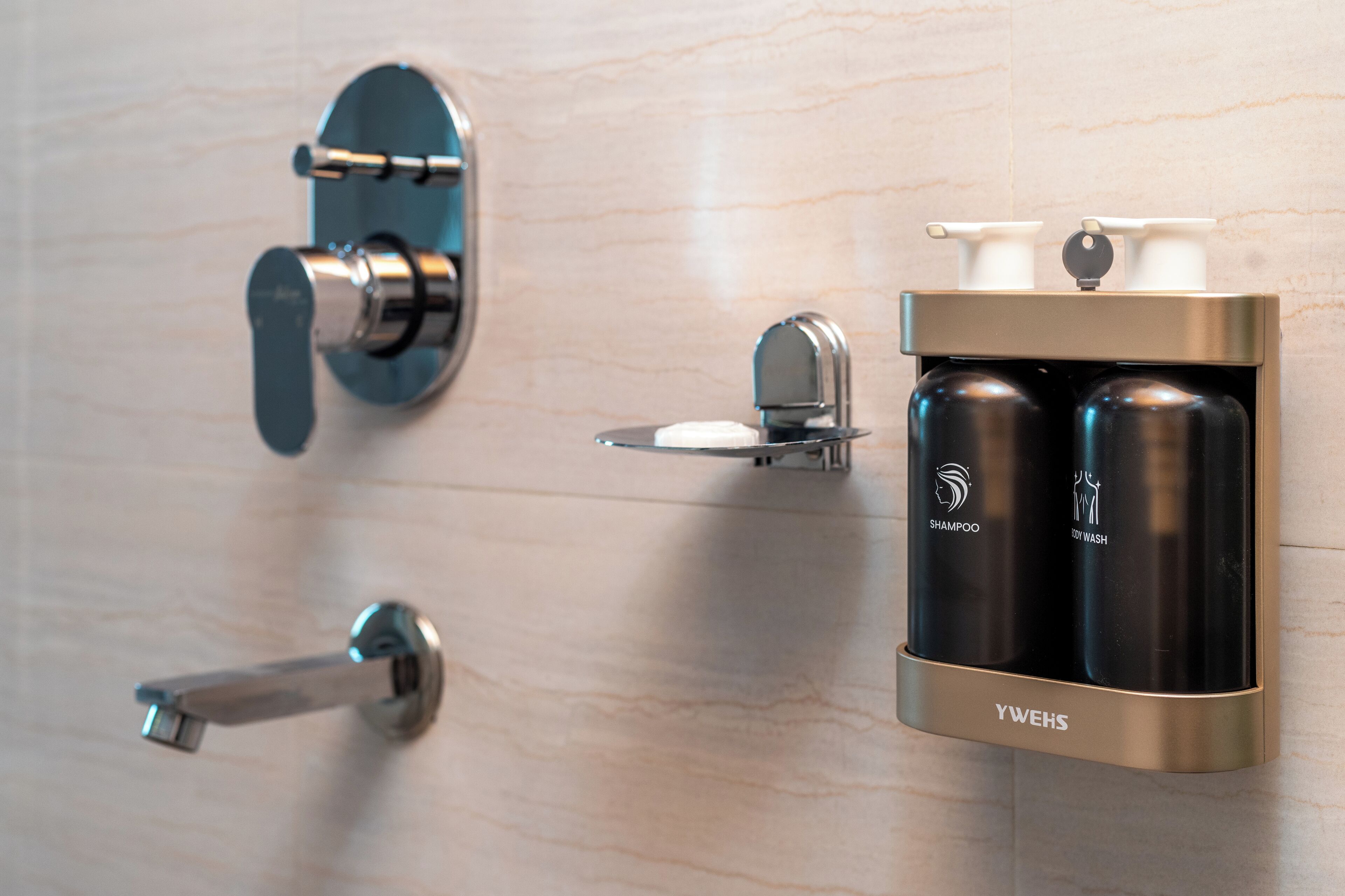 Un baño con un grifo moderno, un dispensador de jabón y dos botellas en la pared, una con la etiqueta 'champú' y otra con la etiqueta 'gel de ducha'.