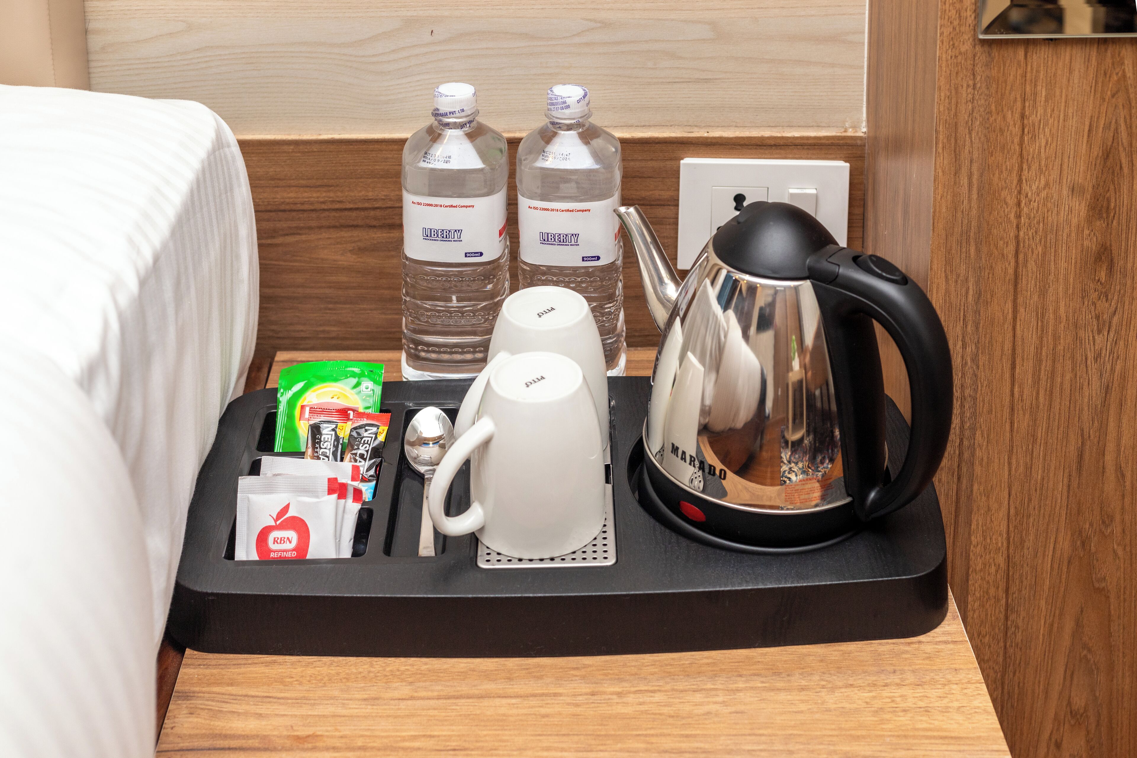 A neatly arranged hotel room with a bed, a tray table with a kettle, two bottles of water, a cup, and some toiletries.