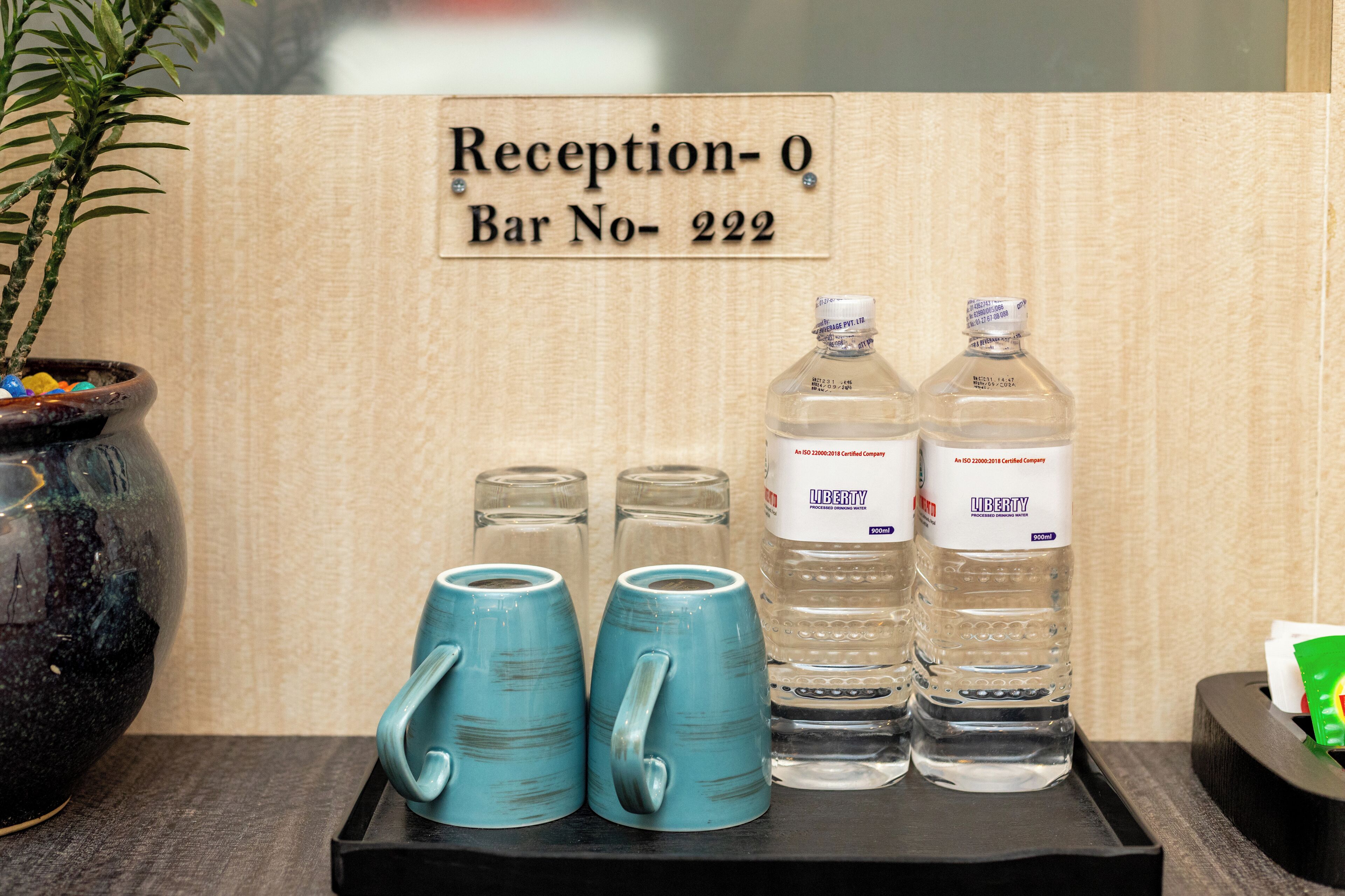 Zona de recepción con un letrero que indica 'Recepción-0 Bar No-222'. Hay dos tazas azules, dos botellas de vidrio transparente etiquetadas 'LIBERTY' y una planta en maceta.