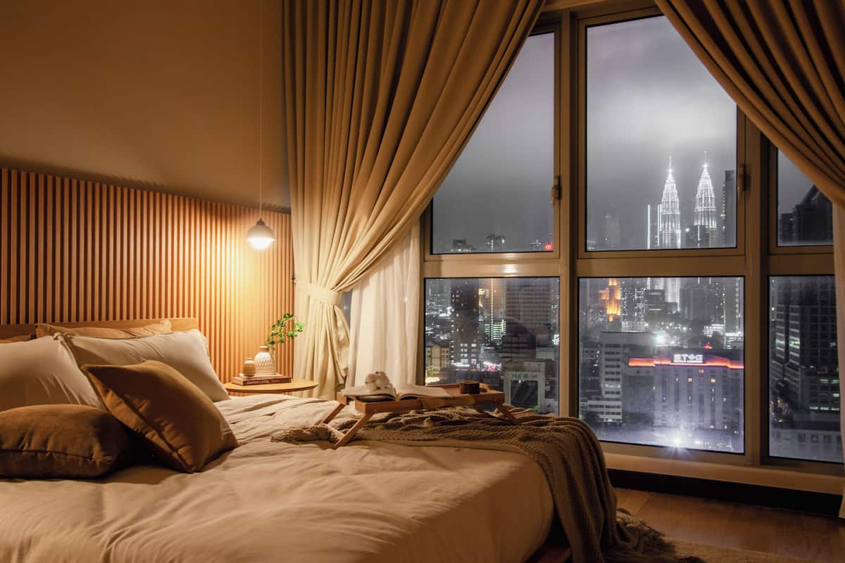 Une chambre avec une grande fenêtre, un lit avec des oreillers, une table de chevet avec une lampe, et une vue sur le paysage urbain la nuit.