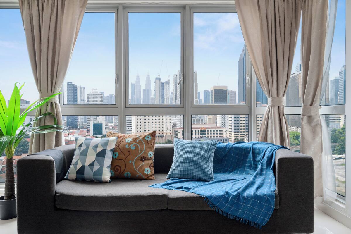 Un salon moderne avec un canapé gris, des coussins à motifs et un plaid bleu. Une grande fenêtre offre une vue sur la ville.