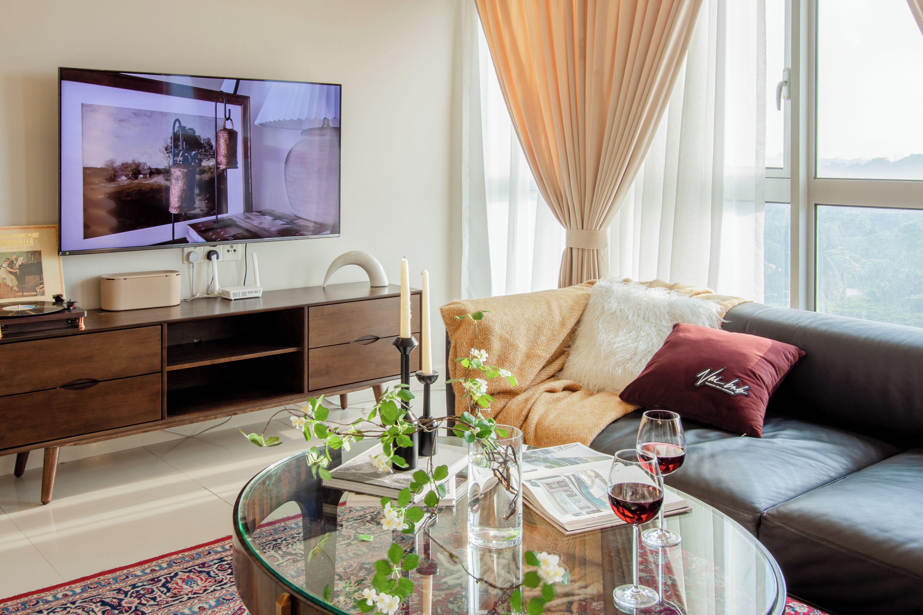 A modern living room with a flat-screen TV, a glass coffee table with flowers, a sofa with cushions, and a view through a window.