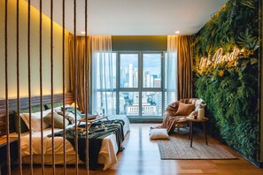 A hotel room with a bed, a chair, a small table, a green wall with a sign, and a large window with a city view.