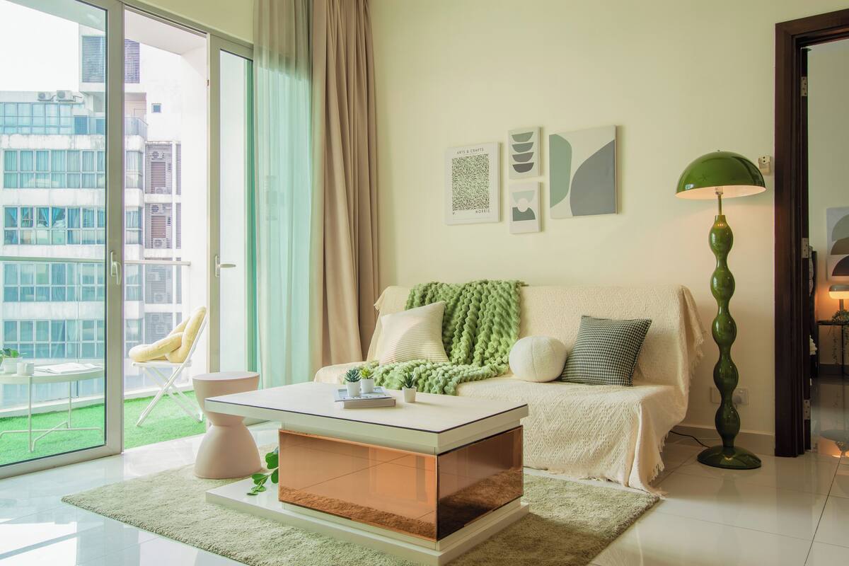 Un salon moderne avec une table basse en verre, un canapé recouvert d’un plaid vert, un lampadaire et une vue sur un immeuble à travers une porte vitrée.