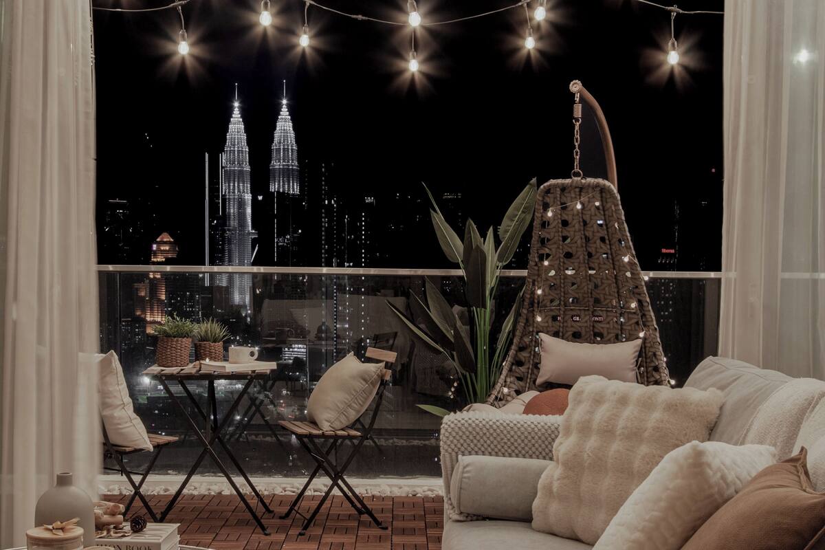 Un balcon offrant une vue sur les tours Petronas la nuit, aménagé avec un canapé, un fauteuil et une chaise suspendue.