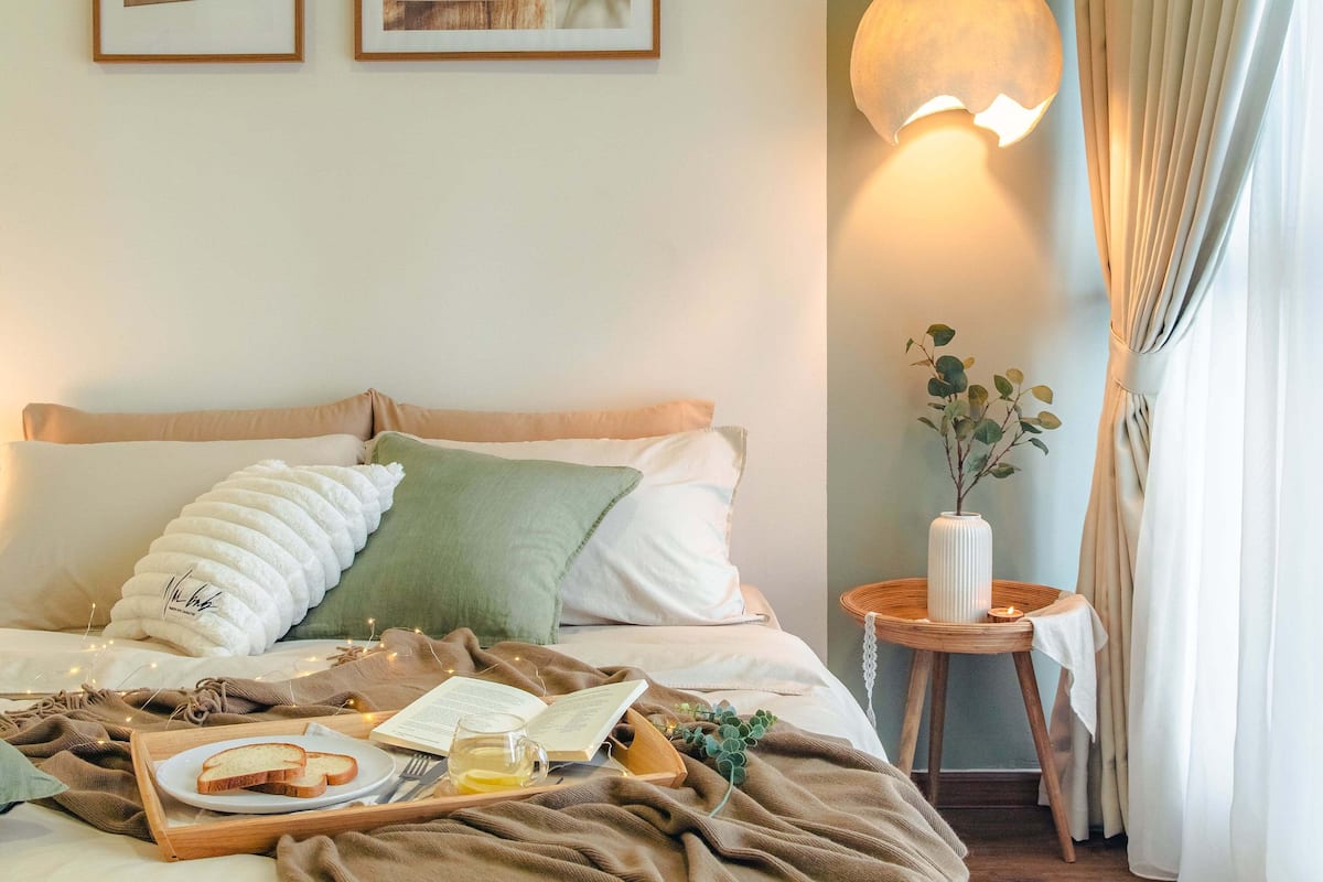 Un lit bien fait, avec des oreillers, une table de chevet ornée d’un vase et d’une bougie, et un plateau contenant du pain et un livre.