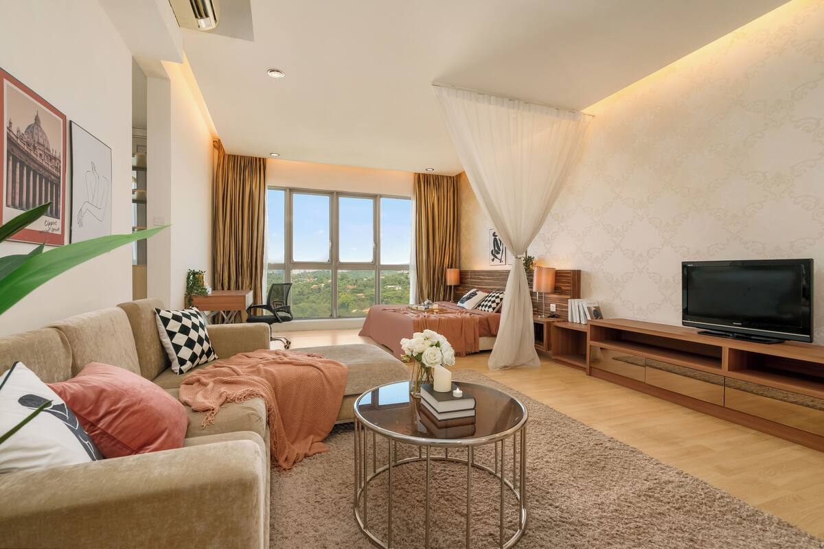 Un salon moderne avec un canapé, une table basse et un téléviseur. Une grande fenêtre, ornée de rideaux, offre une vue sur la verdure environnante.