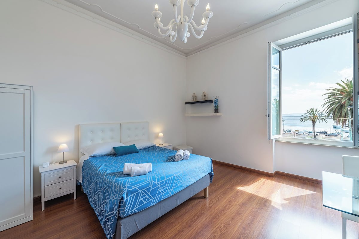 A bedroom with a bed, bedside tables, a nightstand, a chandelier, a window with a view of the sea, and a shelf with decorative items.