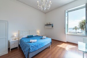 Un dormitorio con cama, mesitas de noche, lámpara de noche, candelabro, ventana con vista al mar y estante con objetos decorativos.
