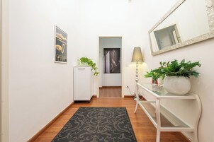 Un pasillo con una puerta blanca, un cuadro en la pared, un armario blanco pequeño, una planta en maceta, una lámpara y una consola con una planta grande.
