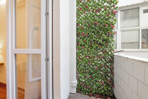A corner of a room with a white door, a window with blinds, and a wall covered with a climbing plant with pink flowers. - Blanco Rooms (Cagliari)