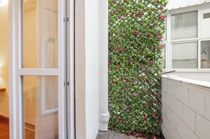 Esquina de una habitación con una puerta blanca, una ventana con persianas y una pared cubierta por una planta trepadora con flores rosas.