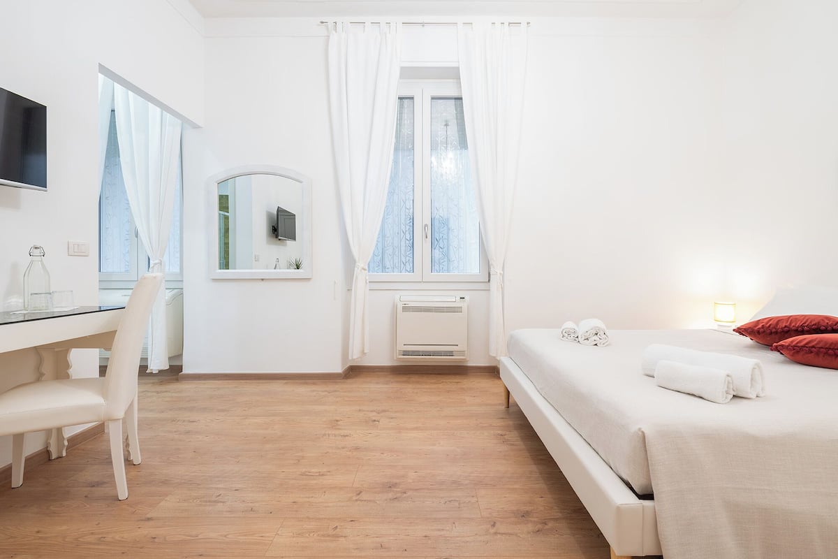 A bedroom with a bed, a desk, a chair, a mirror, and a window with curtains.
