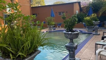 Una piscina con sillas de descanso alrededor y plantas en macetas, frente a un edificio con tejado de baldosas.