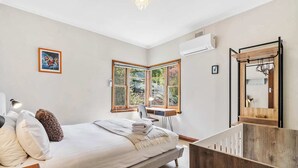 A bedroom with a bed, bedside table, lamp, and a desk with a chair. There is a window with a view of greenery outside. - "Cascade Cottage" South Hobart with parking (South Hobart)