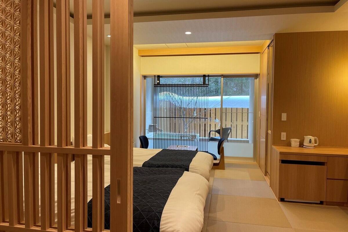 A hotel room with a sliding door, a bed with a black and white bedspread, a small desk with a kettle, and a view of a fenced outdoor area.