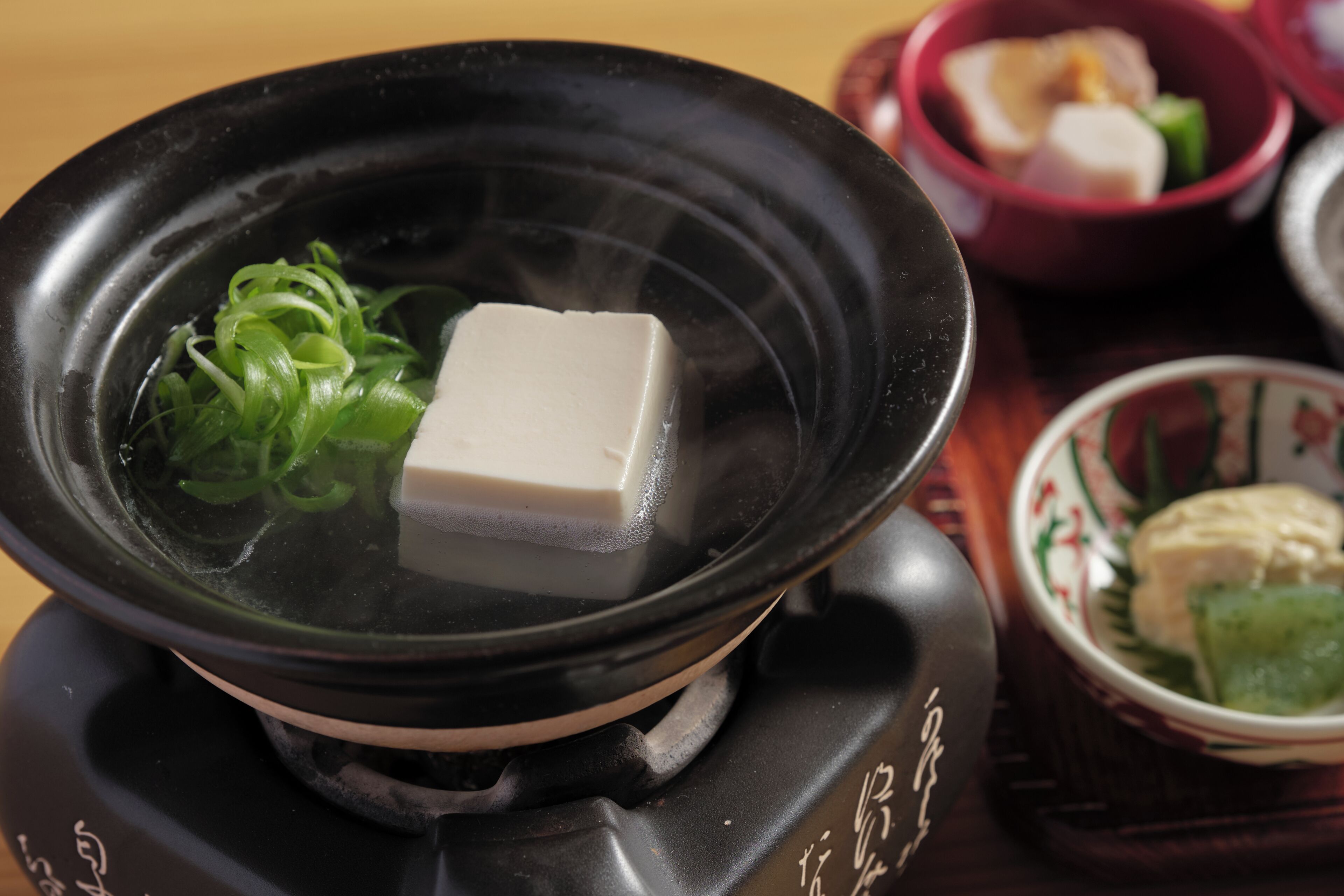 A hot pot with tofu and green onions, a red bowl with food, and a decorative plate with a green leaf.