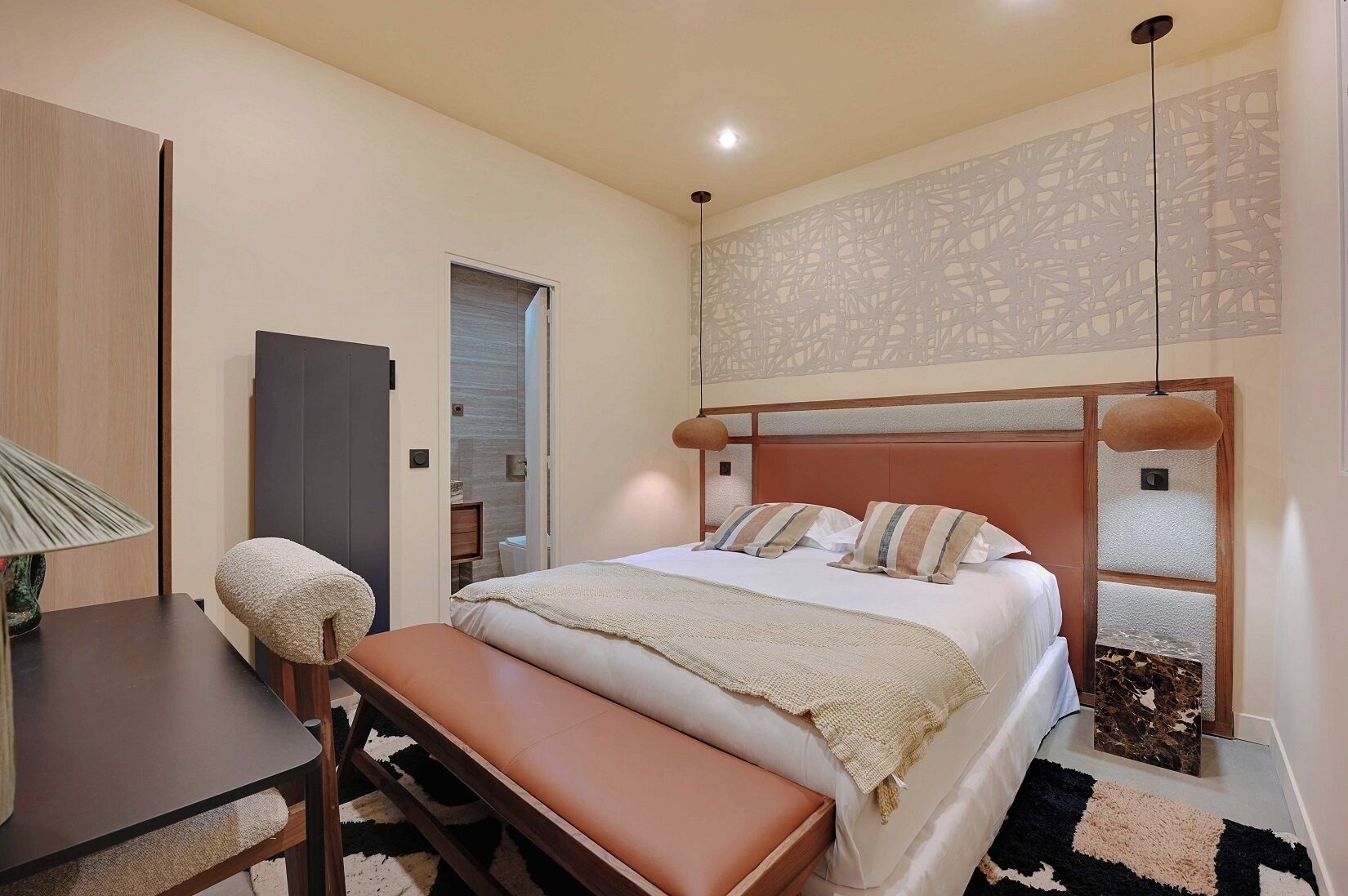 Une chambre à coucher comprenant un lit, un banc, un bureau, une lampe et un panneau décoratif mural.