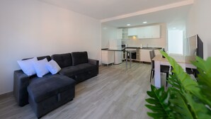 Una sala de estar moderna con un sofá y un reposapiés de color gris oscuro, una mesa de comedor con sillas y una cocina al fondo.