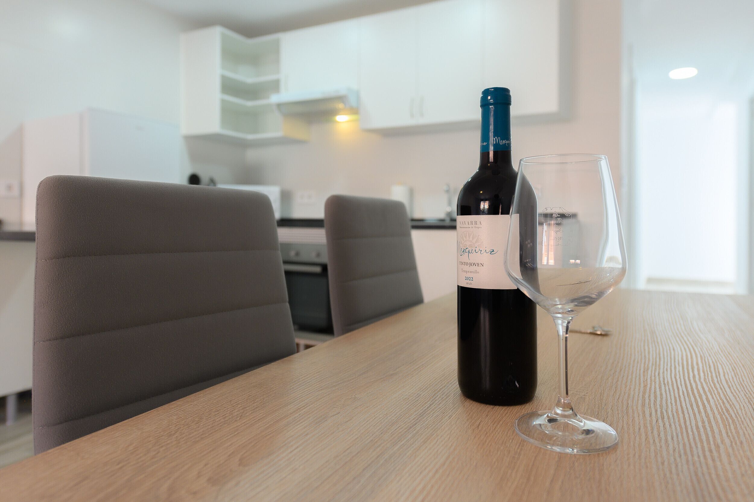 A bottle of wine and an empty glass on a wooden table in a modern kitchen with white cabinets and grey chairs.