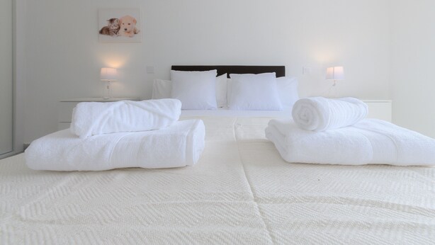 Una cama bien hecha con sábanas y almohadas blancas, una mesita de noche con lámpara y un cuadro en la pared.