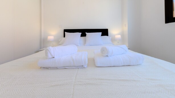 Una cama bien tendida con sábanas y almohadas blancas, dos lámparas de noche y una habitación limpia y de color blanco.