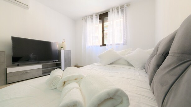 Een netjes opgemaakt bed met witte lakens en een grijs hoofdeinde, een flatscreen televisie op een houten standaard en een raam met transparante gordijnen.