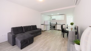 Een moderne woonkamer met een grijze hoekbank, een eethoek met een glazen tafel, en een keuken op de achtergrond.