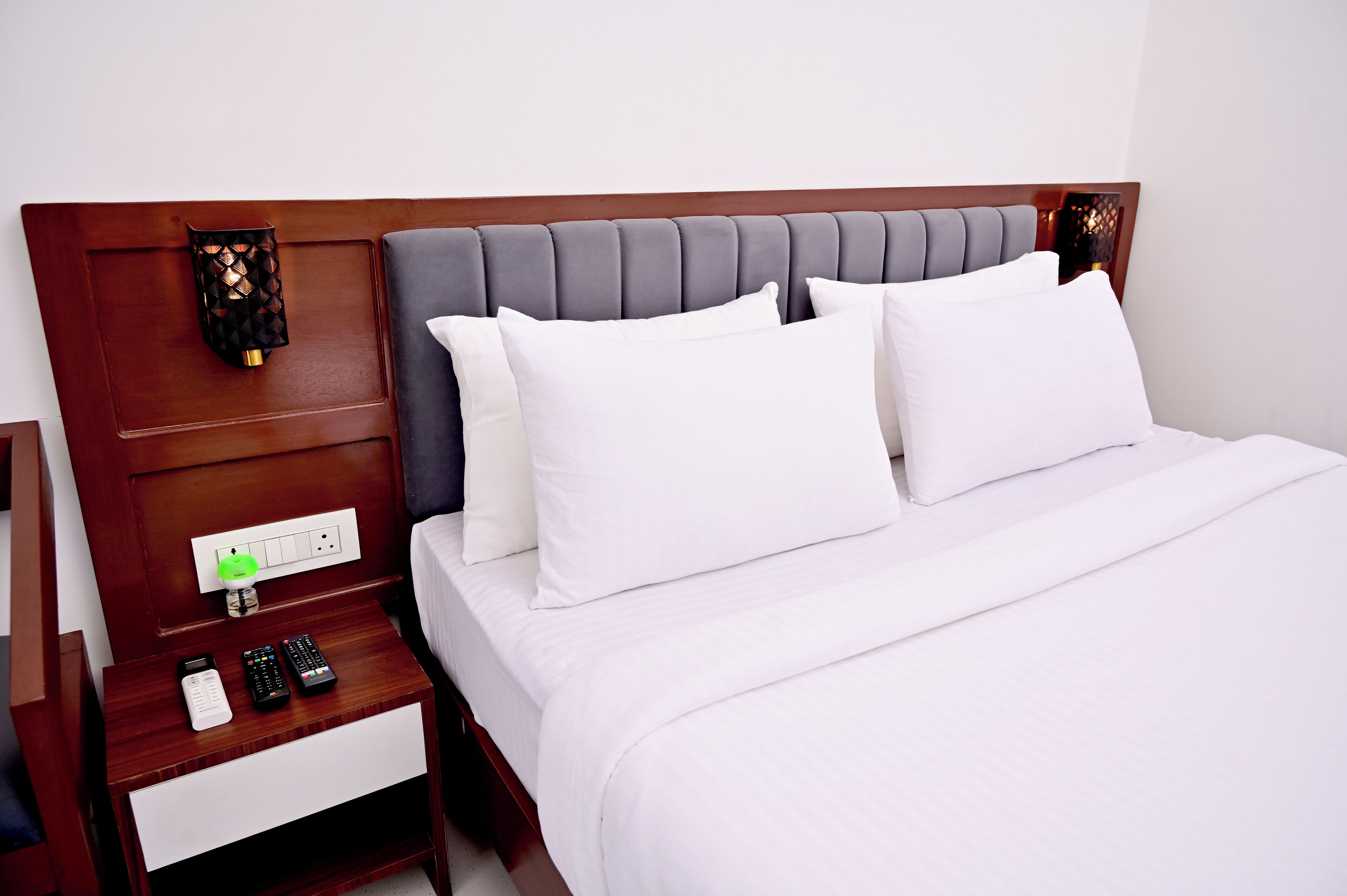 A neatly made bed with white linens, a wooden headboard, and a nightstand with remote controls and a power outlet.