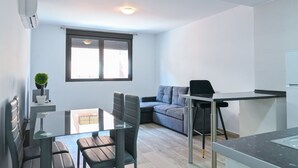 Un espacio habitable moderno y compacto con zona de comedor, un sofá gris, una pequeña cocina y un ventanal que proporciona luz natural.