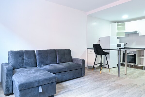 A modern living room with a grey sectional sofa and a black chair, a kitchen with white cabinets and stainless steel appliances, and a light wood floor.