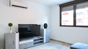 Una sala de estar moderna con un televisor de pantalla plana, un mueble gris, una planta pequeña y una ventana con persianas.