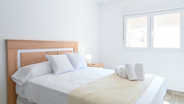 Una cama bien hecha con sábanas blancas y un cabecero de madera, una mesita de noche con una lámpara, y una ventana con persianas.