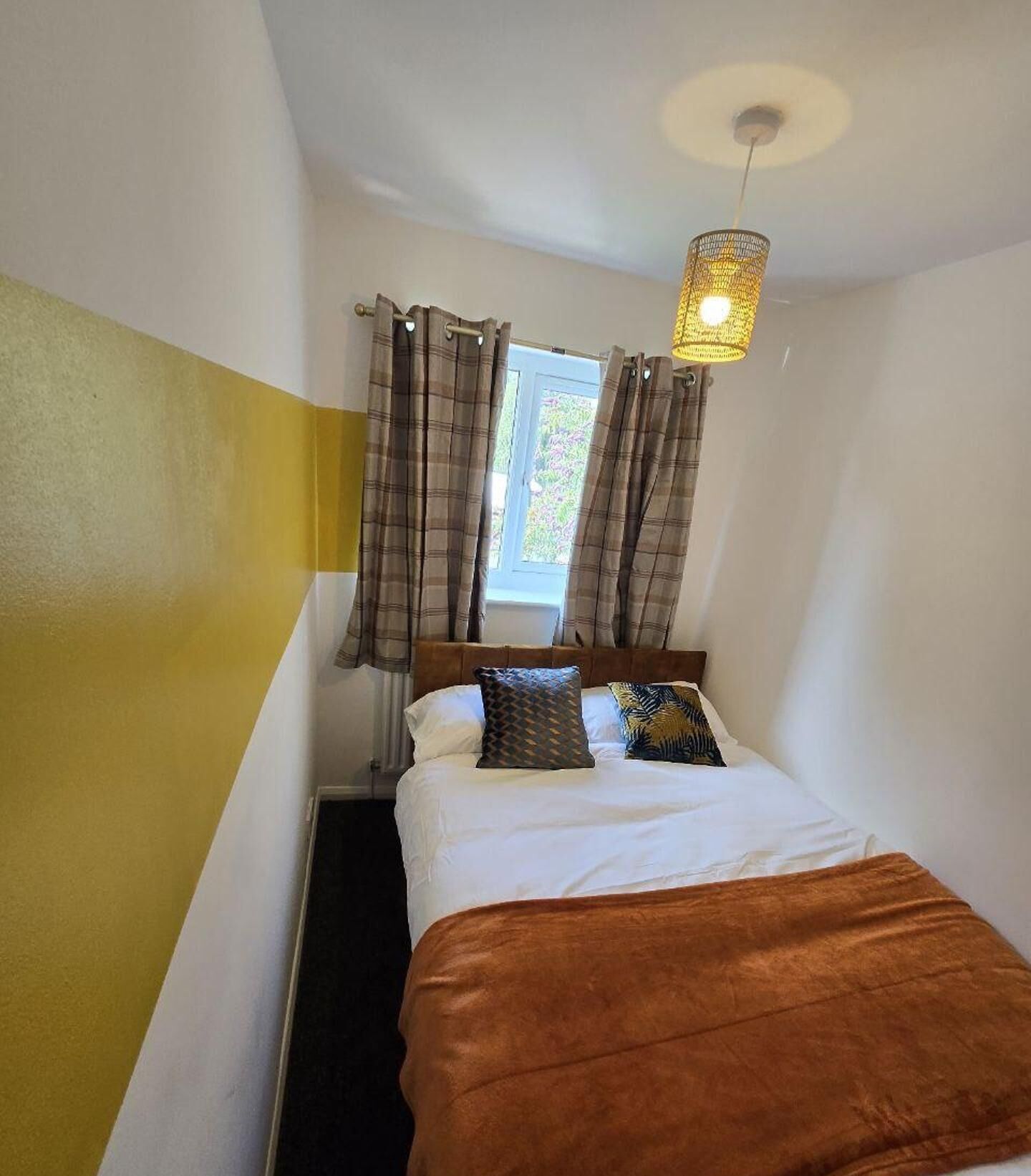 A narrow room with a bed, a window with curtains, and a hanging light fixture.