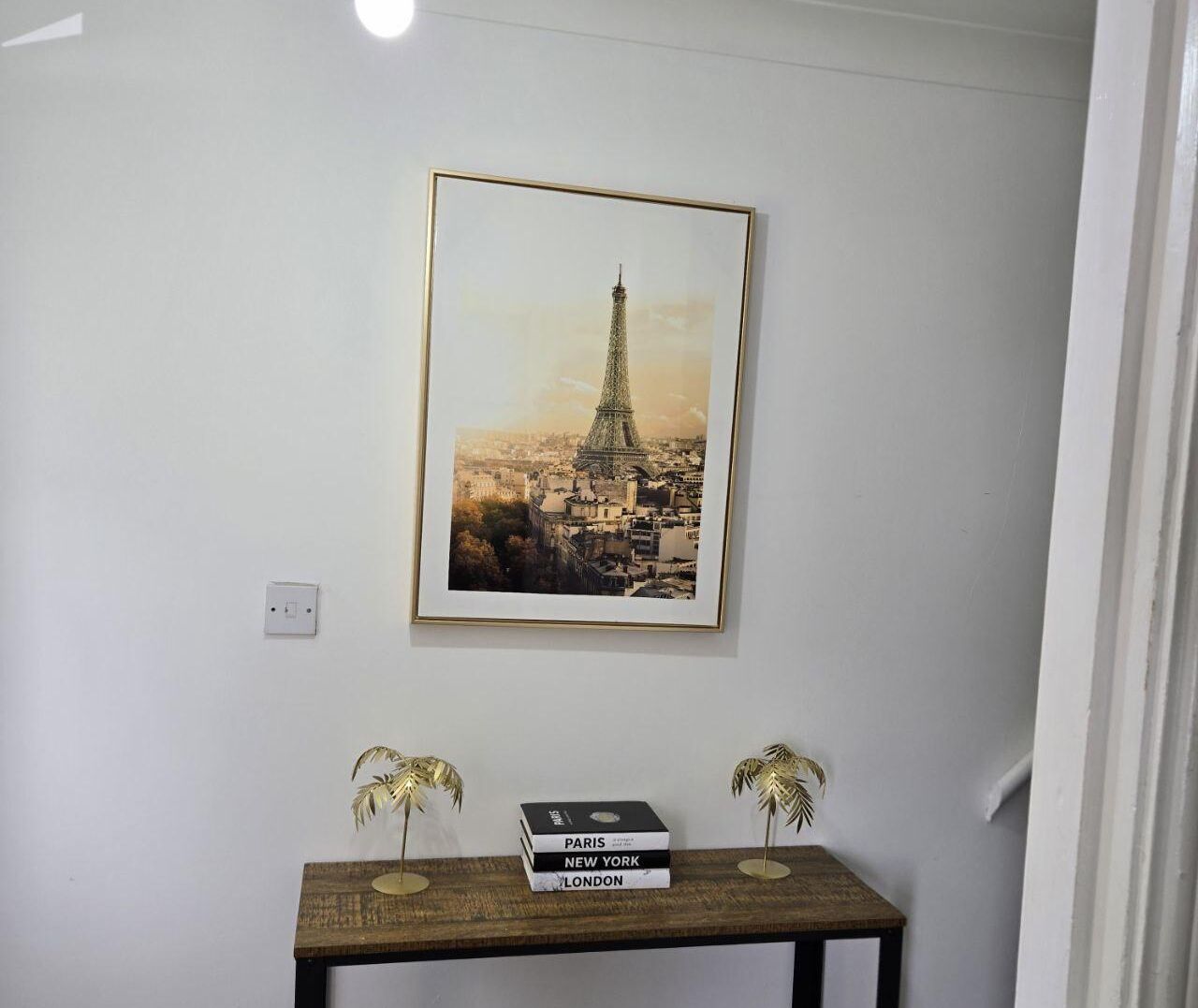 A framed picture of the Eiffel Tower on a wall, a small wooden table with books and two decorative items, and a light switch.