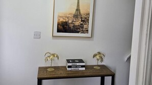 Un tableau encadré représentant la Tour Eiffel, accroché au mur, une petite table en bois avec des livres et deux objets décoratifs, et un interrupteur.