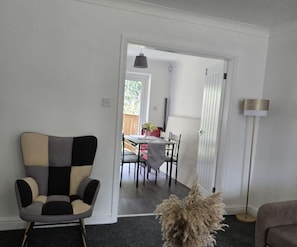 A living room with a modern chair, a small table with chairs, a floor lamp, and a doorway leading to another room.