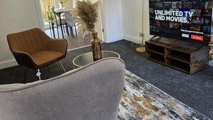 Un salon avec un canapé gris, un téléviseur diffusant une offre de « télévision et de films à volonté », un grand lampadaire et un tapis à motifs.