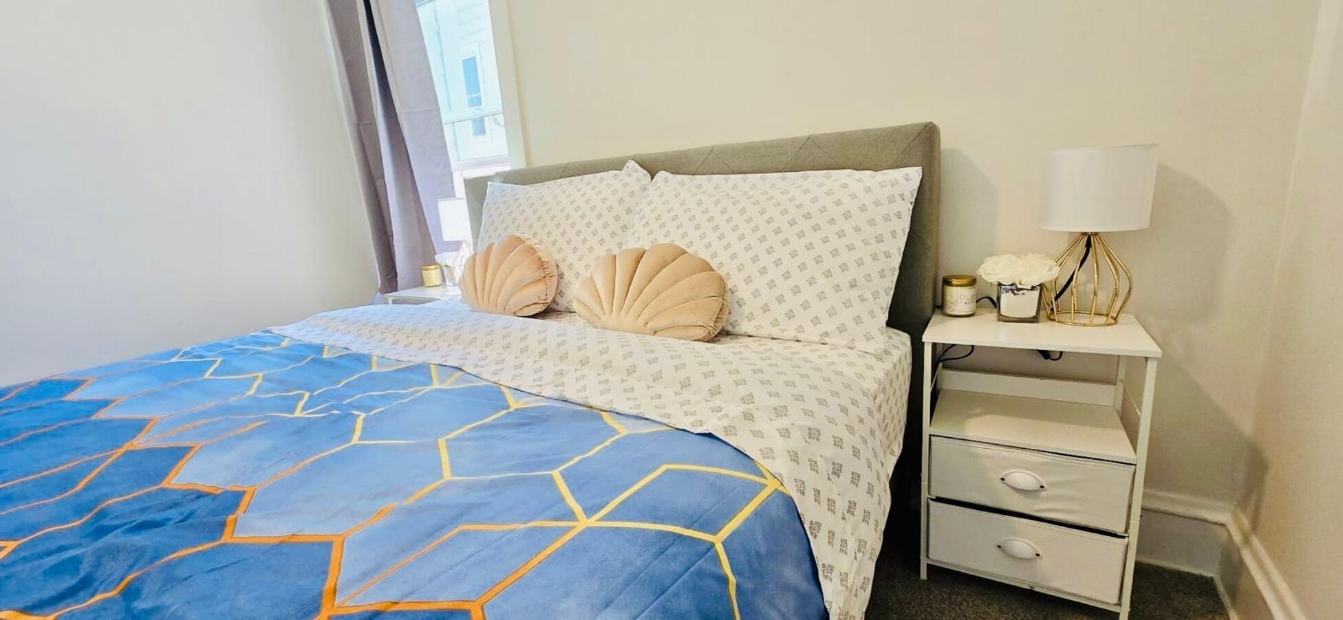 Un lit bien fait, recouvert d’une couvre-lit aux motifs géométriques bleus et jaunes, avec une tête de lit rembourrée grise et deux oreillers beige. On aperçoit une table de chevet blanche, sur laquelle se trouvent une lampe, un petit objet décoratif et un récipient. La pièce a des murs blancs et une fenêtre donnant sur des bâtiments à l’extérieur.