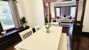 A dining area with a white table and chairs, a vase with flowers, and a view of a living room with a sofa and a window. - 2024 Remodeled 2bedroom in West CLE (Cleveland)