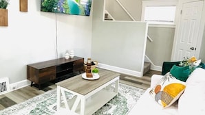 A living room with a white sofa, a coffee table, a flat-screen TV, and a staircase leading to an upper level. - Olive and Sunshine CLE House (Cleveland)