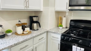 A kitchen with white cabinets, a granite countertop, a black stove, and various kitchen items like a coffee maker, jars, and a small plant. - Olive and Sunshine CLE House (Cleveland)