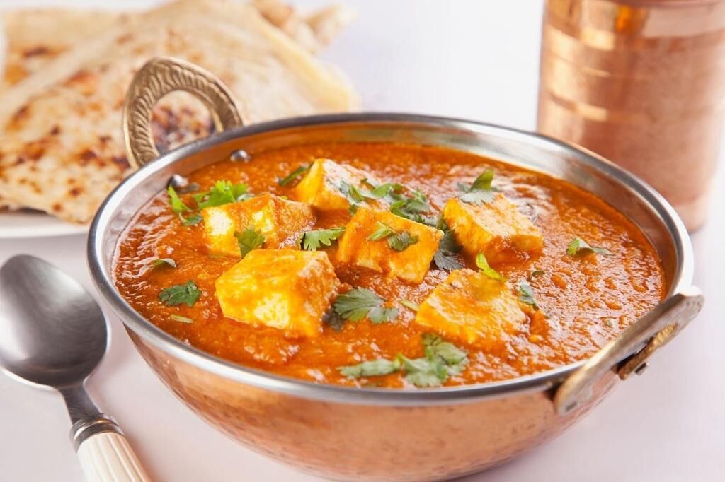 A bowl of paneer curry garnished with cilantro, accompanied by naan bread and a glass of beverage.