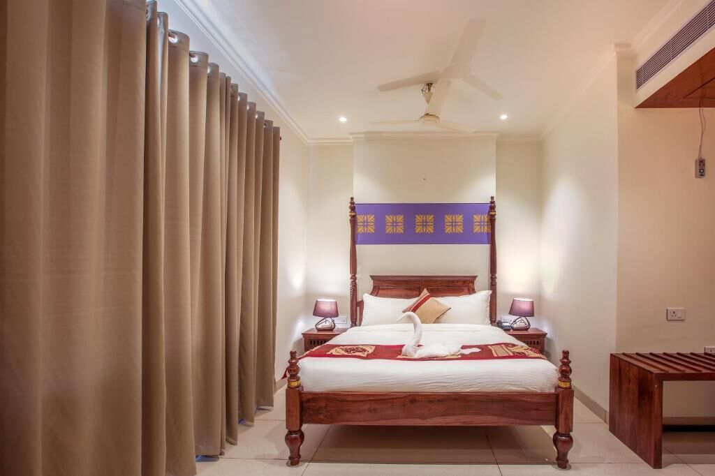 A hotel room with a wooden four-poster bed, beige curtains, a ceiling fan, and a purple decorative panel above the bed.